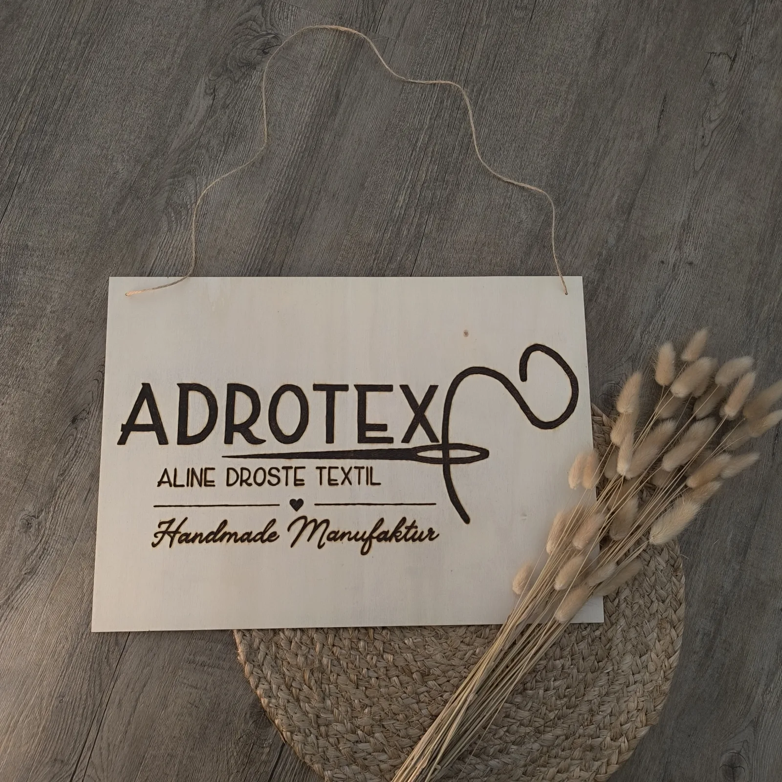 Graviertes Holzschild mit Jutekordel, ADROTEX Logo und Trockenblumen daneben.