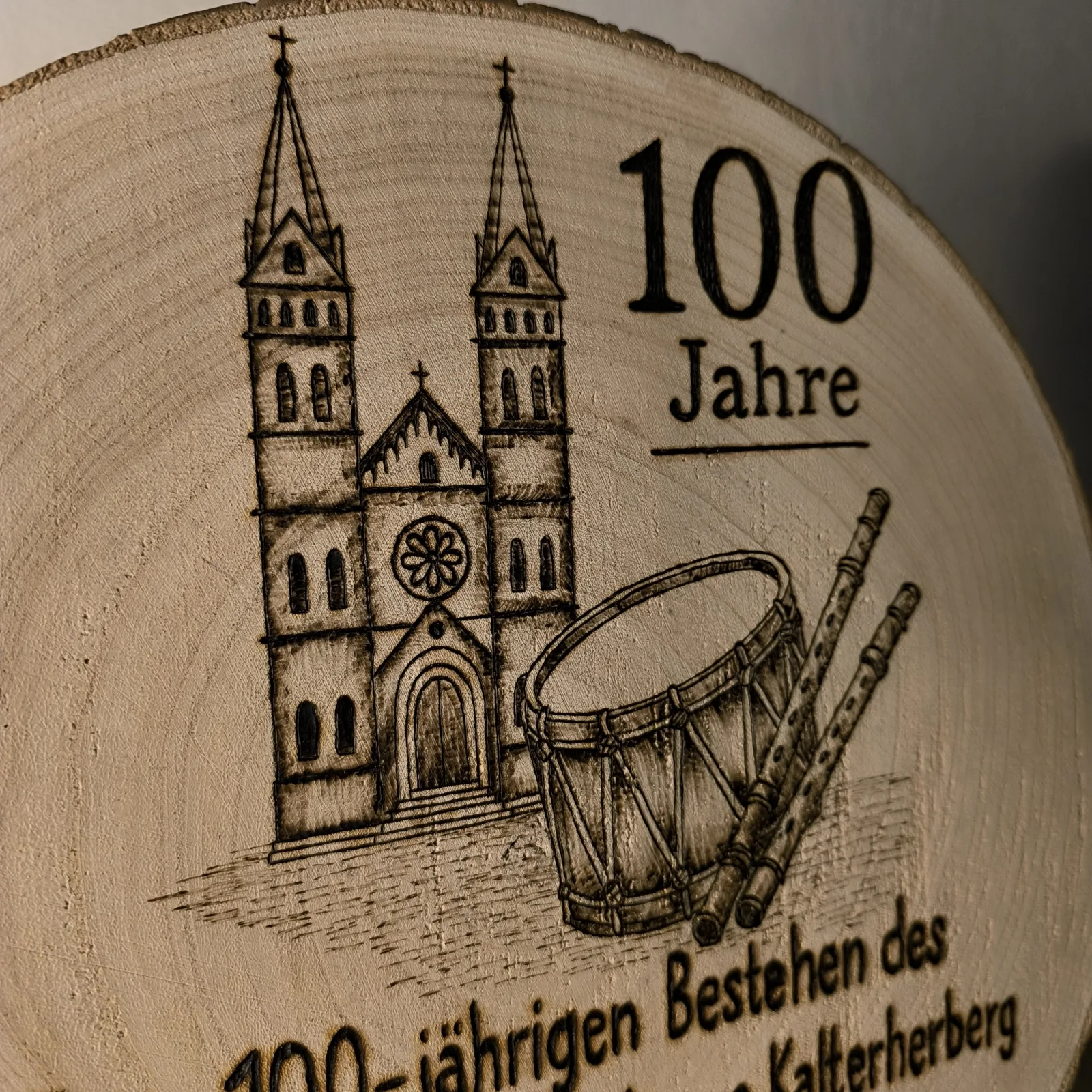 Nahaufnahme der Holzscheibe mit Kirchenmotiv, Trommel, Pfeifen und Aufschrift 100 Jahre.