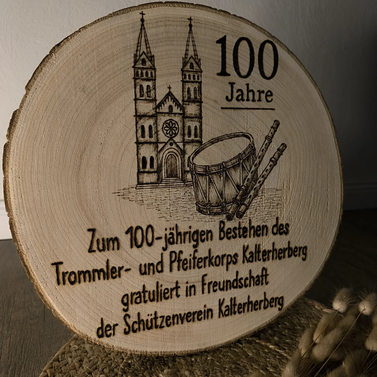 Runde Holzscheibe mit Kirche, Trommel und Pfeifen sowie Aufschrift 100 Jahre.
