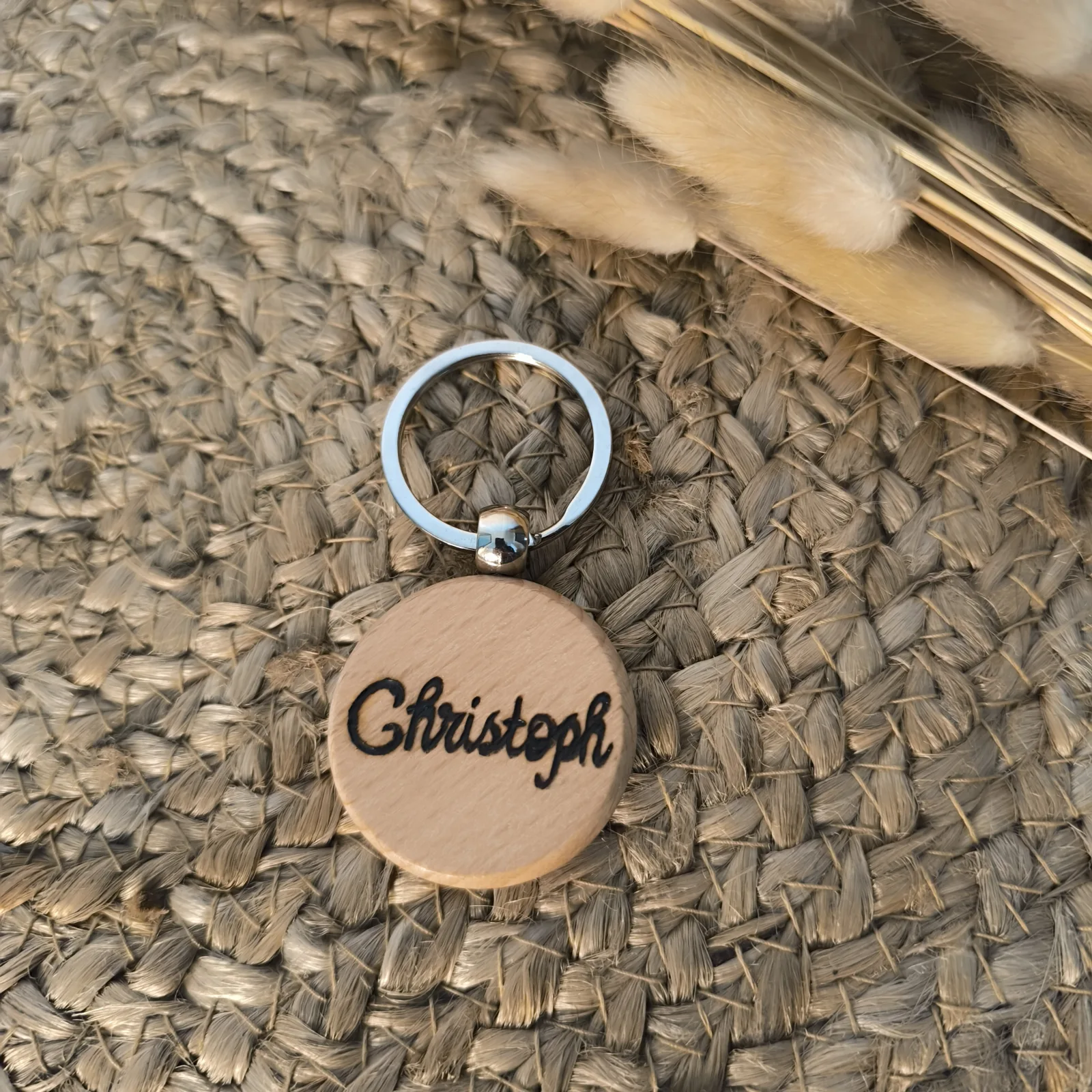 Runder Holzschlüsselanhänger mit Metallring und Aufschrift Christoph neben Trockenblumen.
