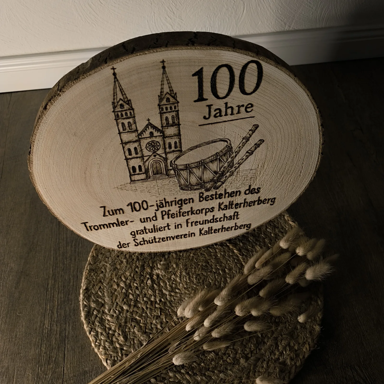 Schräg stehende Holzscheibe mit Brandmalerei zu 100 Jahren und Musikmotiv.